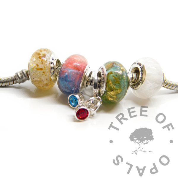 engraved bead quad, umbilical cord bead, cord stump in resin with unicorn white resin sparkle mix and dual colour breastmilk bead, dragon's blood red and aegean blue resin sparkle mix. Birthstone dangle charm duo on European dangle charm setting. Slim European setting with July and September birthstones, dual colour umbilical cord bead, basiliak green and aegean blue resin sparkle mix with lots of gold filled leaf, breastmilk bead with very subtle coloured pearlescent shimmer, shown on European style snake chain bracelet. Solid sterling silver bead cores. Engraving in arial font